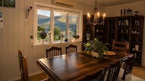 A dining room with a wooden table, chairs, a chandelier, a bookshelf, and a window with a view of mountains. - Room-casa Hauge B&B Sunrise Room (Gloppen)