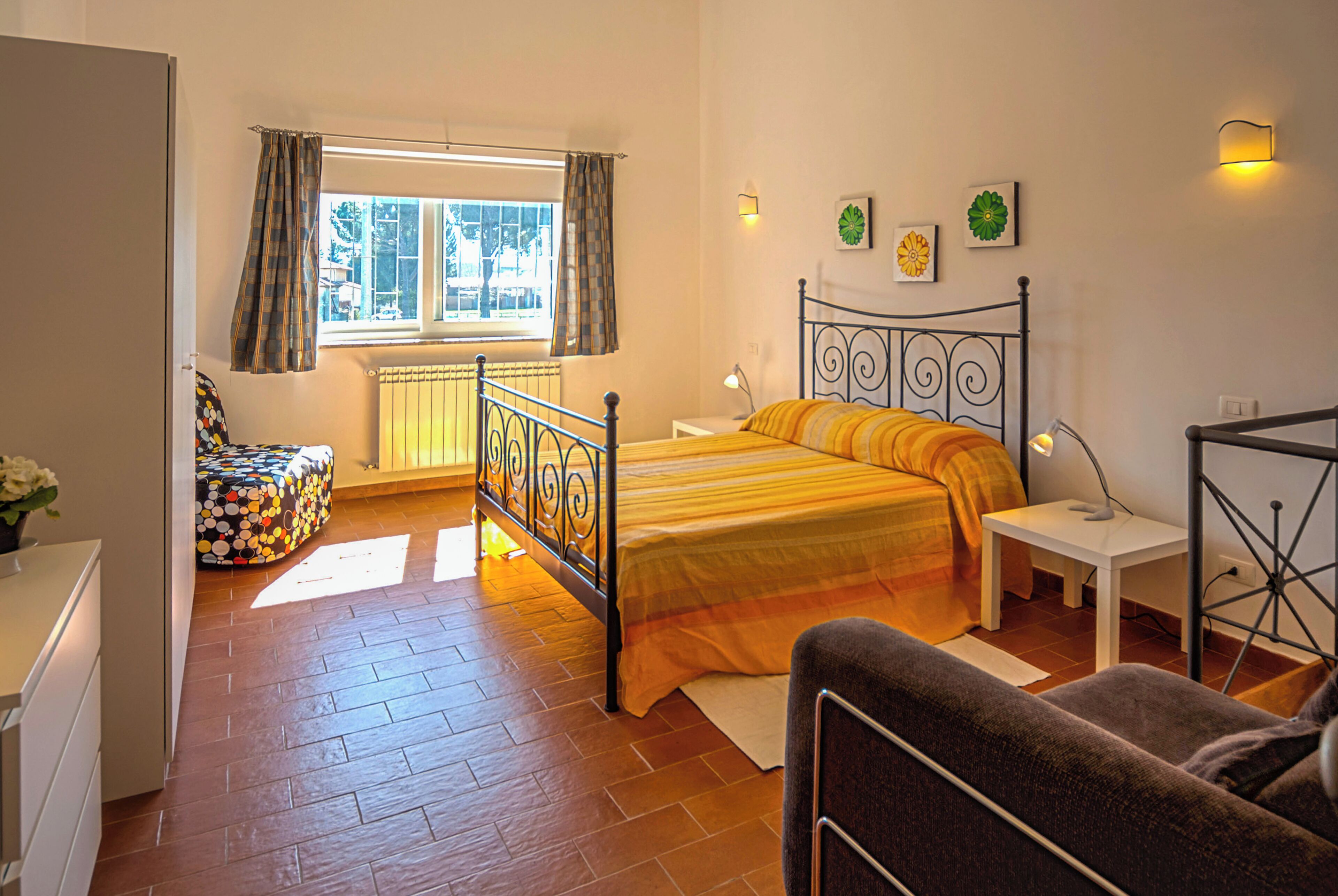 A bedroom with a bed, a chair, a bedside table, a lamp, a window with curtains, and a radiator.