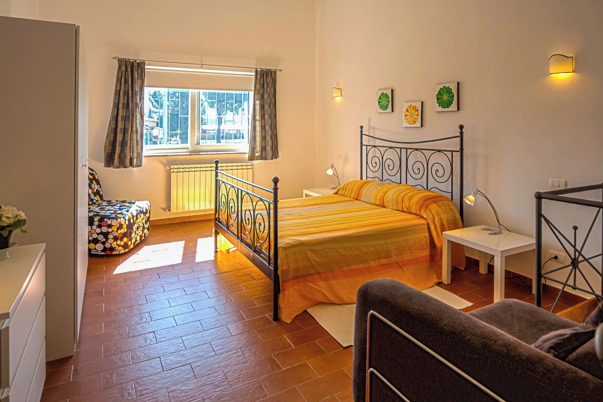 A bedroom with a bed, a chair, a bedside table, a lamp, a window with curtains, and a radiator.