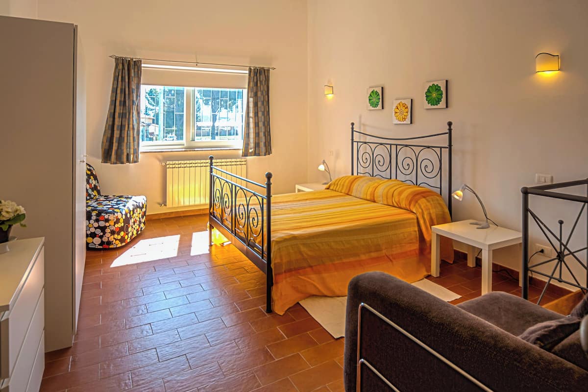 A bedroom with a bed, a chair, a bedside table, a lamp, a window with curtains, and a radiator.