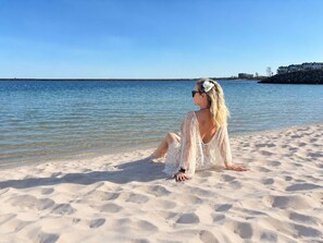 Eine Person sitzt an einem sandigen Strand mit freiem Blick auf das Meer und eine entfernte Küste.