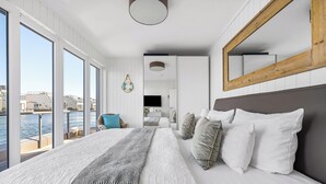 Ein modernes Schlafzimmer mit einem großen Bett, Blick auf das Wasser und einem spiegelnden Wandbild mit Holzrahmen.