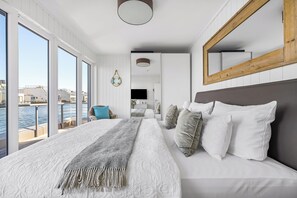 Ein modernes Schlafzimmer mit einem großen Bett, Blick auf das Wasser und einem spiegelnden Wandbild mit Holzrahmen.