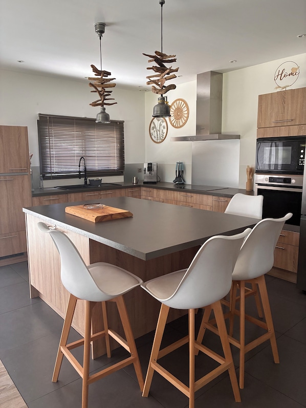 A modern kitchen with a large island, wooden bar stools, a coffee machine, and a microwave.