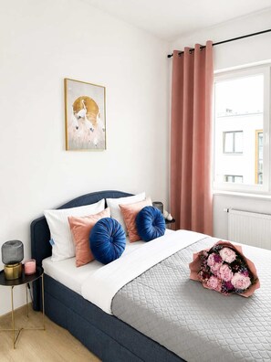Un dormitorio con cama, almohadas, mesita de noche, lámpara, una obra de arte en la pared, ventana con cortinas y un ramo de flores.