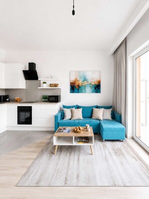 A modern living room with a blue sofa, a coffee table, and a kitchen area with white cabinets and a black range hood. - Lumina Apart near ZOO in Lodz with Parking (Łódź)