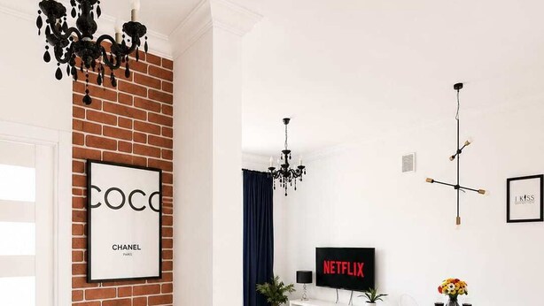 Ein modernes Wohnzimmer mit einer Ziegelwand, einem Netflix-Schild, einer Kristallleuchtre und einem Essbereich mit weißem Tisch und Stühlen.