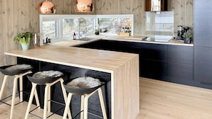 A modern kitchen with wooden panel walls, a central island, bar stools, pendant lights, and a view through large windows. - Architect drawn home with amazing view over Nisser (Treungen)