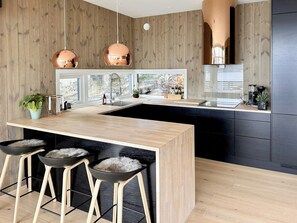 A modern kitchen with wooden panel walls, a central island, bar stools, pendant lights, and a view through large windows. - Architect drawn home with amazing view over Nisser (Treungen)