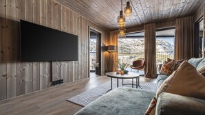Ein modernes Wohnzimmer mit Flachbildfernseher, Holzwänden, einem runden Couchtisch, einer Couch und Blick auf eine gebirgige Landschaft.