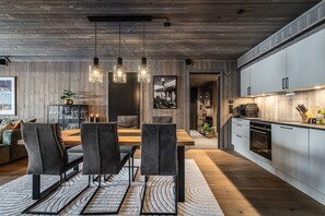 Cuina moderna amb armaris de fusta, zona de menjador amb taula i cadires de fusta, i lluminàries penjants.