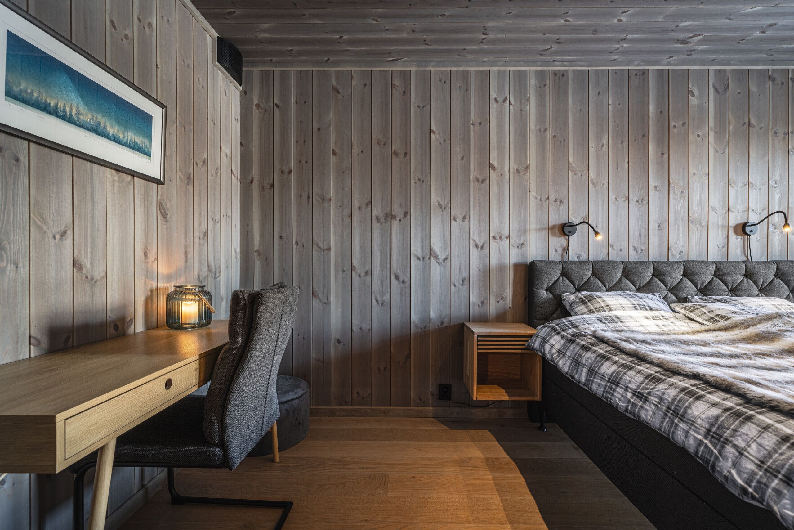 Um quarto com parede de madeira, uma cama com cabeceira estofada, uma escrivaninha com cadeira e uma mesa de cabeceira com abajur.