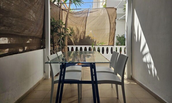 Mesa de vidrio con sillas en un espacio exterior iluminado por el sol, con una pared blanca y una zona con sombra.