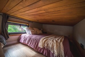 Une chambre avec un plafond en bois, un lit recouvert d’une courtepointe rouge, une fenêtre avec des rideaux et une vue sur la verdure à l’extérieur.