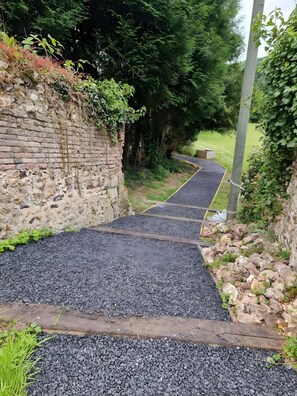 A gravel pathway leading through a natural area with a brick wall and greenery on one side.