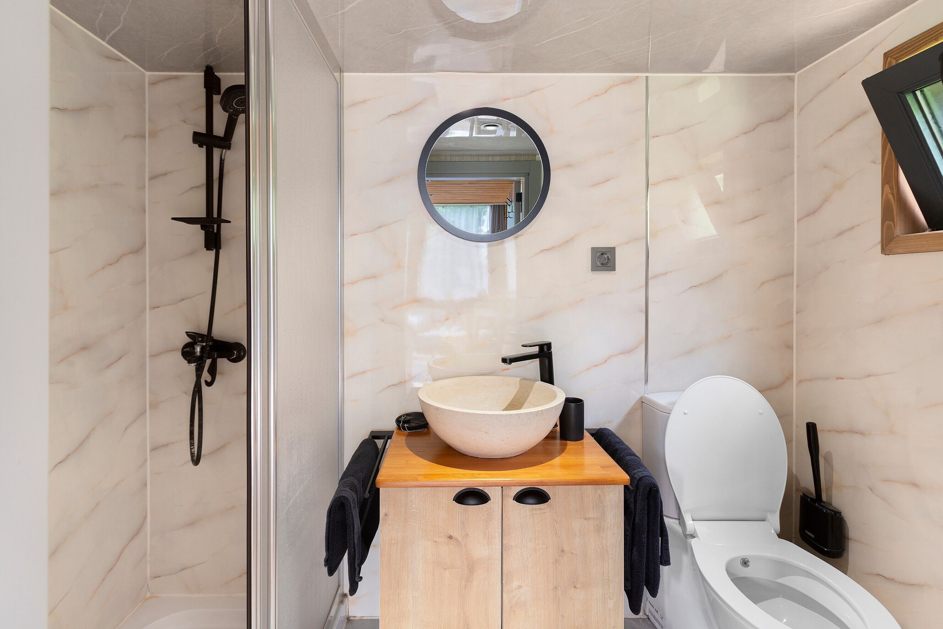 A bathroom with a wooden vanity, a round mirror, a white sink, a toilet, and a shower area.