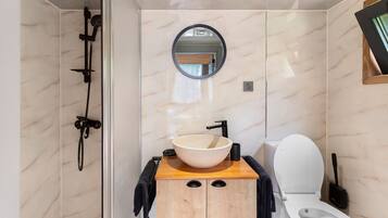 A bathroom with a wooden vanity, a round mirror, a white sink, a toilet, and a shower area.