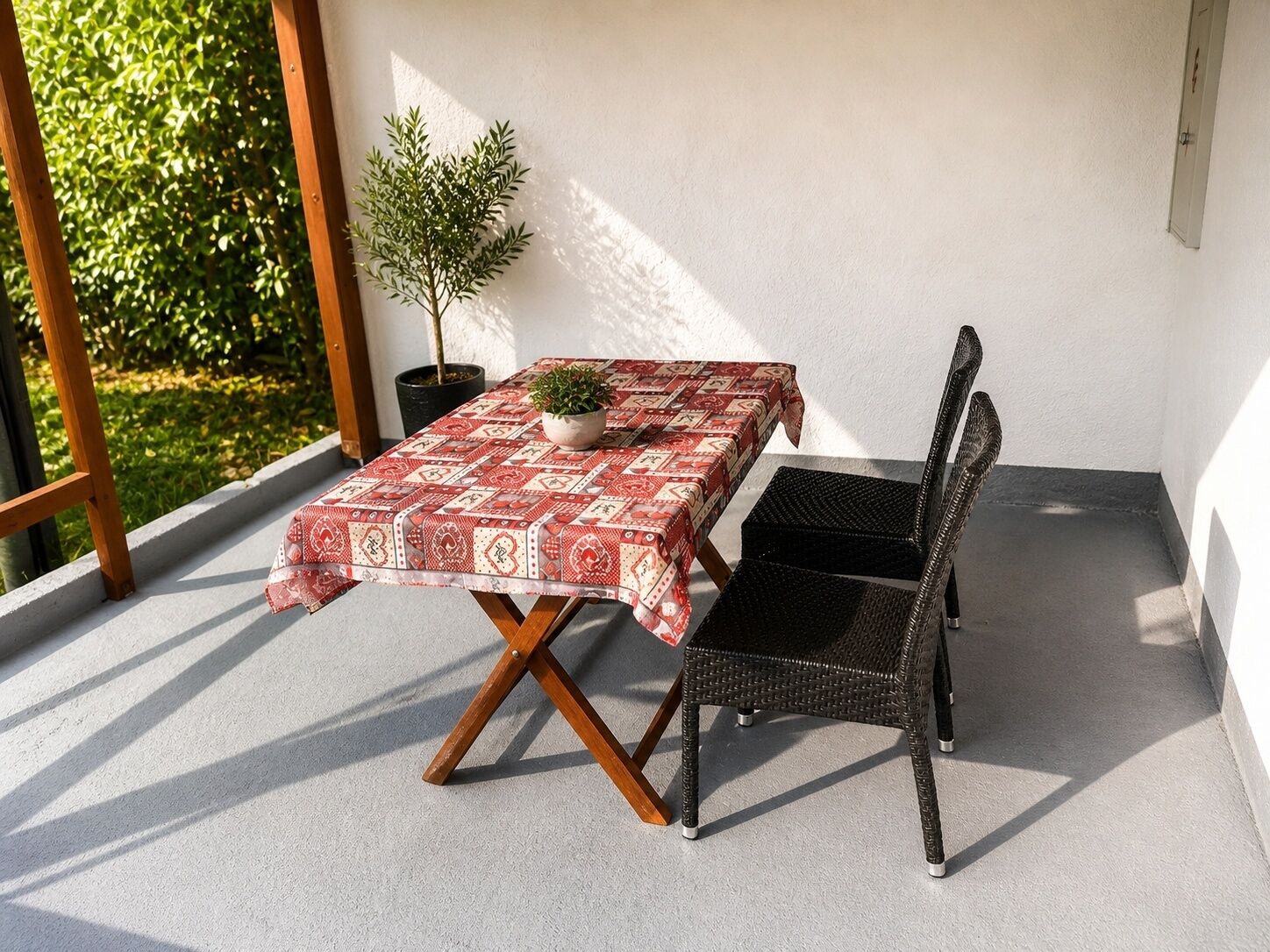 An outdoor patio with a table covered in a patterned tablecloth, a potted plant, and a wicker chair.