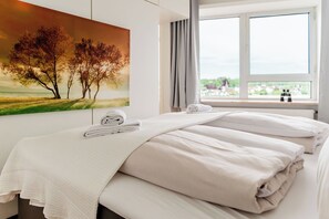 A hotel room with a bed, a painting of trees, a window with a view, and a towel on the bed. - Vacation apartment "FjordHavn" on the 8th floor of the Viking Tower (Schleswig)