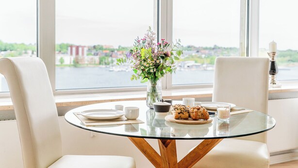 Een ronde glazen tafel met houten poten, omringd door witte stoelen, een vaas met bloemen, borden, bekers en een glas melk.
