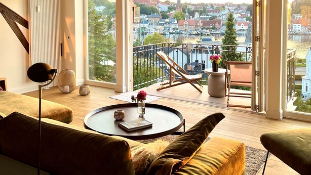 A modern living room with a large window, a round coffee table, a sofa, and a balcony with a view of a cityscape. - "Hafenpanorama" vacation rental with a view of the Old Town and the harbor, located right in (Flensburg)