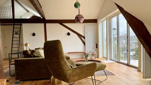 A loft-style room with a high ceiling, wooden beams, a green armchair, a sofa, a ladder, and large windows with a city view. - "Hafenpanorama" vacation rental with a view of the Old Town and the harbor, located right in (Flensburg)