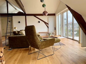 A loft-style room with a high ceiling, wooden beams, a green armchair, a sofa, a ladder, and large windows with a city view. - "Hafenpanorama" vacation rental with a view of the Old Town and the harbor, located right in (Flensburg)