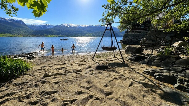 Una playa arenosa con un columpio, un banco de madera y un edificio de piedra con vistas a montañas y un lago.