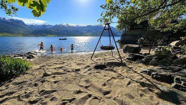 Une plage de sable avec une balançoire, un banc en bois et un bâtiment en pierre offrant une vue sur les montagnes et un lac.