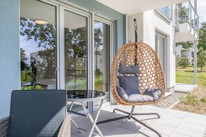 Ein Korbstuhl mit Kissen, ein Glastisch und ein Sessel ebenfalls mit Kissen auf einer Terrasse mit Glastür und Blick auf Grünflächen.