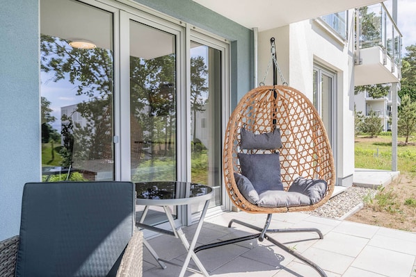 Ein Korbstuhl mit Kissen, ein Glastisch und ein Sessel ebenfalls mit Kissen auf einer Terrasse mit Glastür und Blick auf Grünflächen.