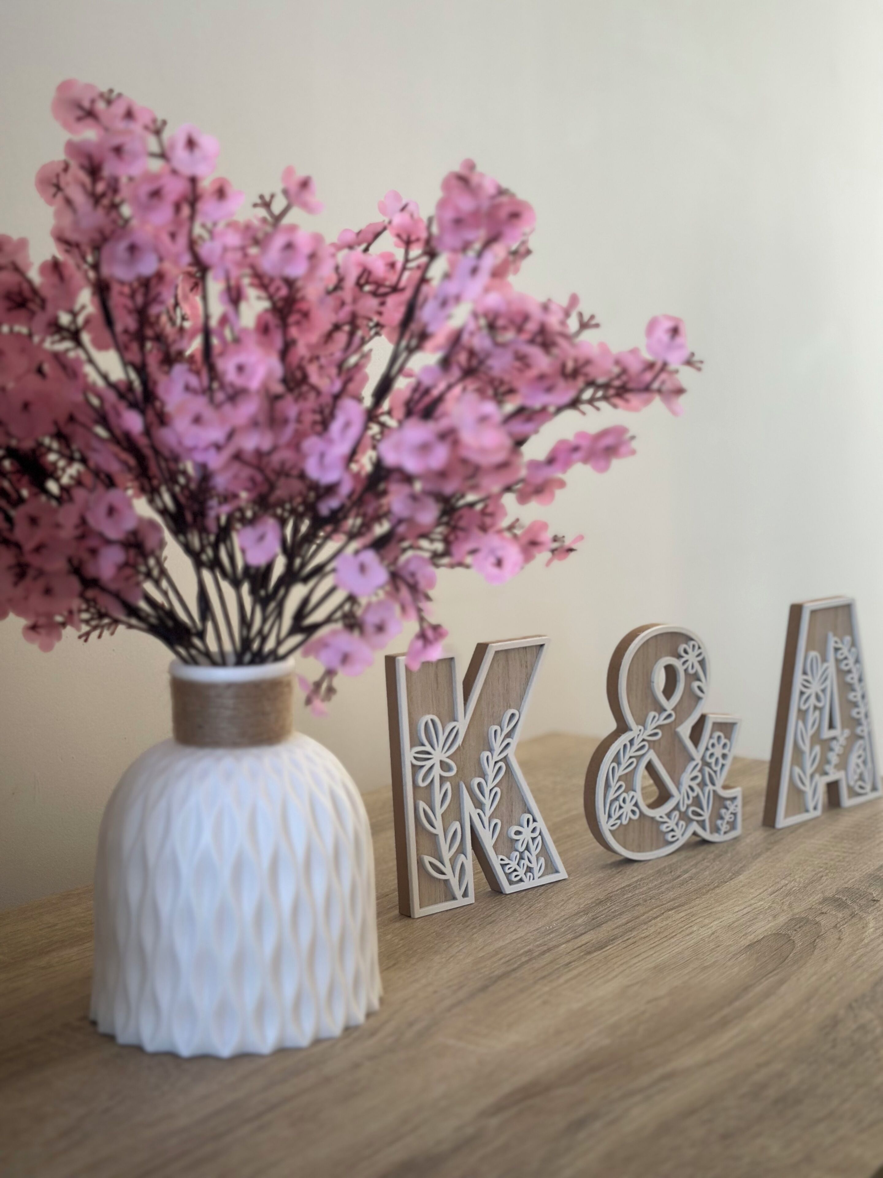 En vit vas med rosa blommor och träbokstäver som bildar orden ”K & A”, placerad på en träyta.