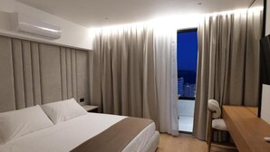 A hotel room with a bed, a desk, a chair, a TV, and a view of the city at night. - Safir Luxury Suites (Sarandë)