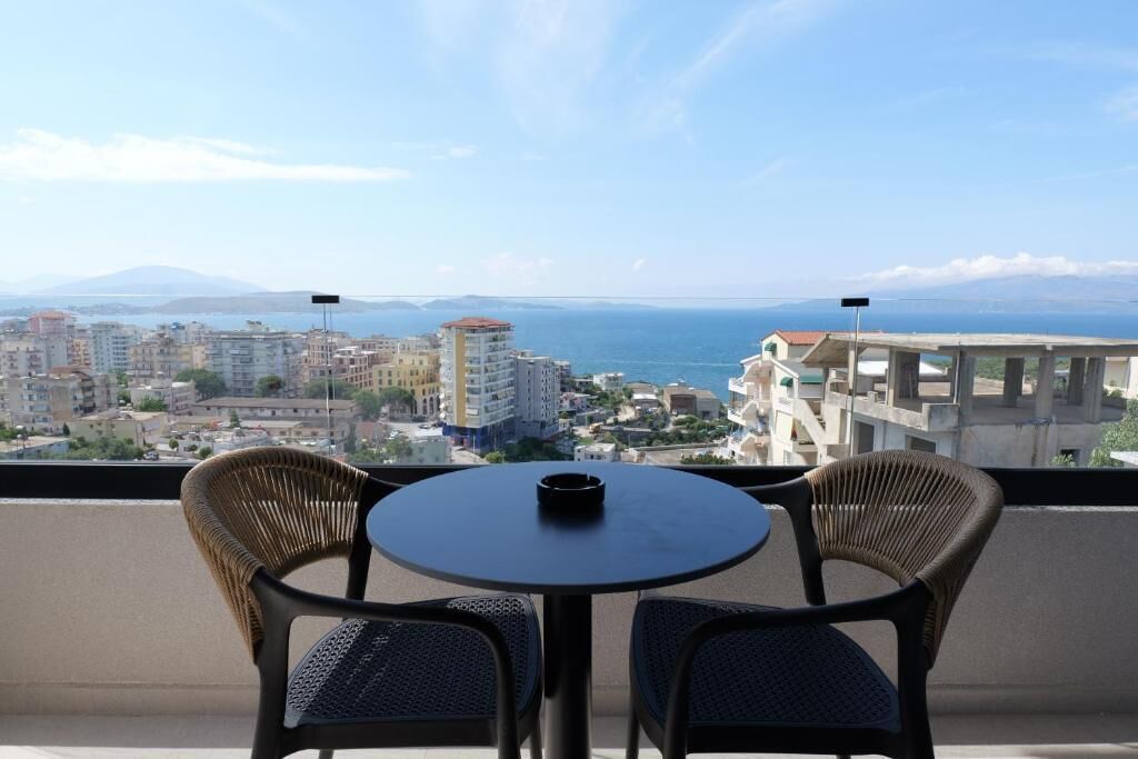 A balcony with wicker chairs and a table, overlooking a coastal cityscape with buildings and the sea.