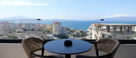 A balcony with wicker chairs and a table, overlooking a coastal cityscape with buildings and the sea.