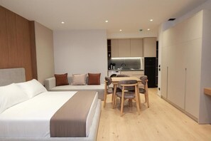 A modern hotel room with a bed, a seating area with a table and chairs, a kitchenette, and a wardrobe.