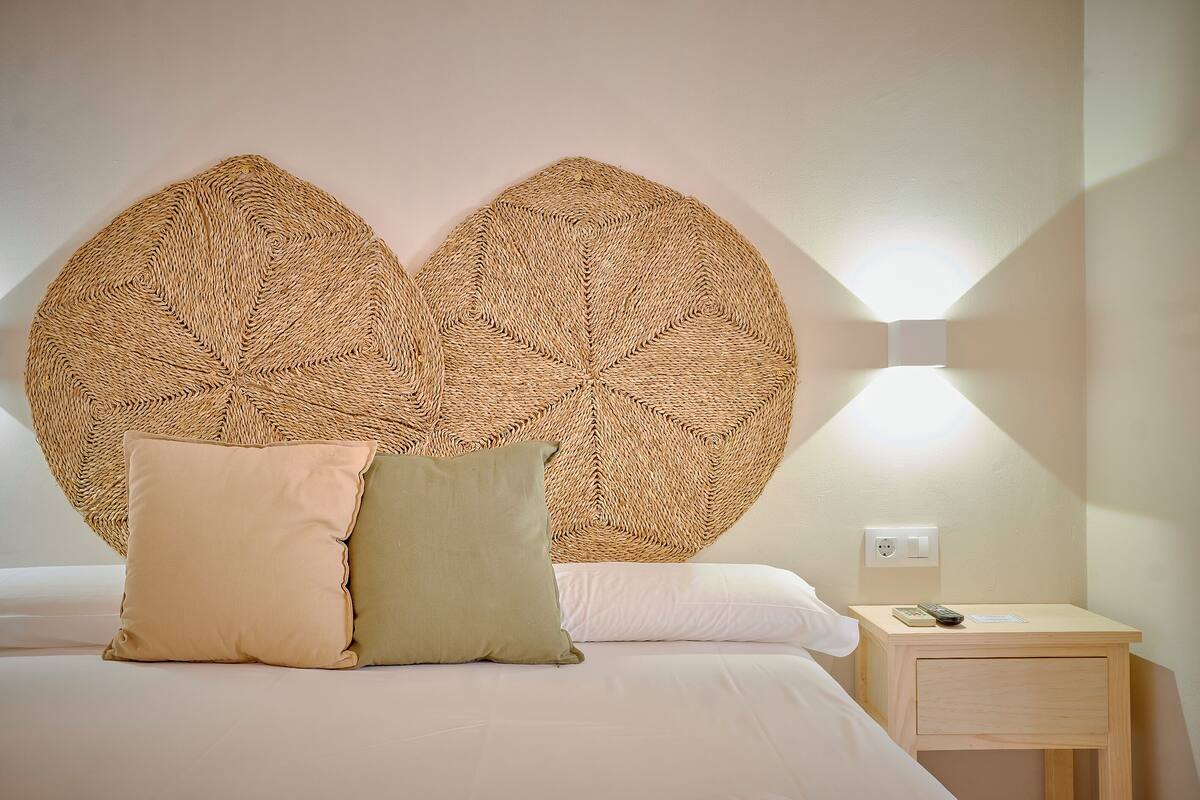 A bed with a woven headboard, two pillows, a bedside table with a remote control, and a wall socket.