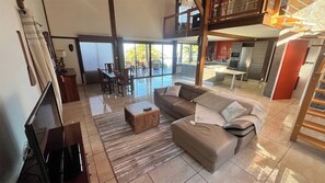 A spacious living area with a large sofa, a coffee table, and a TV. There is a balcony with a view of the outdoors. - Villa TheOceane (Les Avirons)