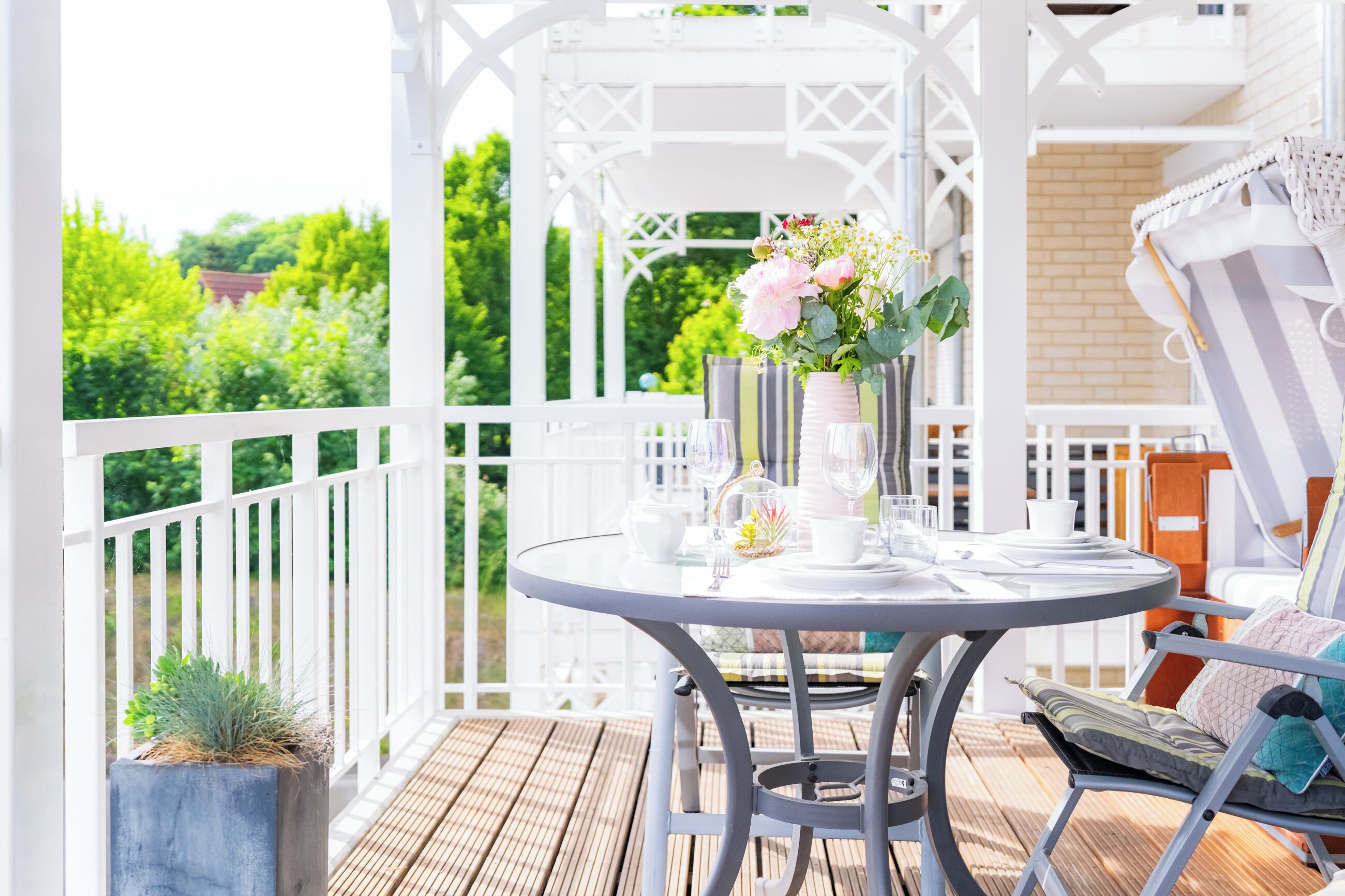 A covered outdoor patio with a round table set for dining, surrounded by white railings and a view of greenery.