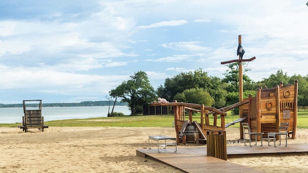 Una estructura de juego de madera con toboganes y elementos para escalar, una terraza de madera y una silla de playa.