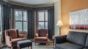 Ein Eckzimmer mit zwei Sesseln, einem Sofa, einer Lampe und einem Fenster mit Vorhängen.