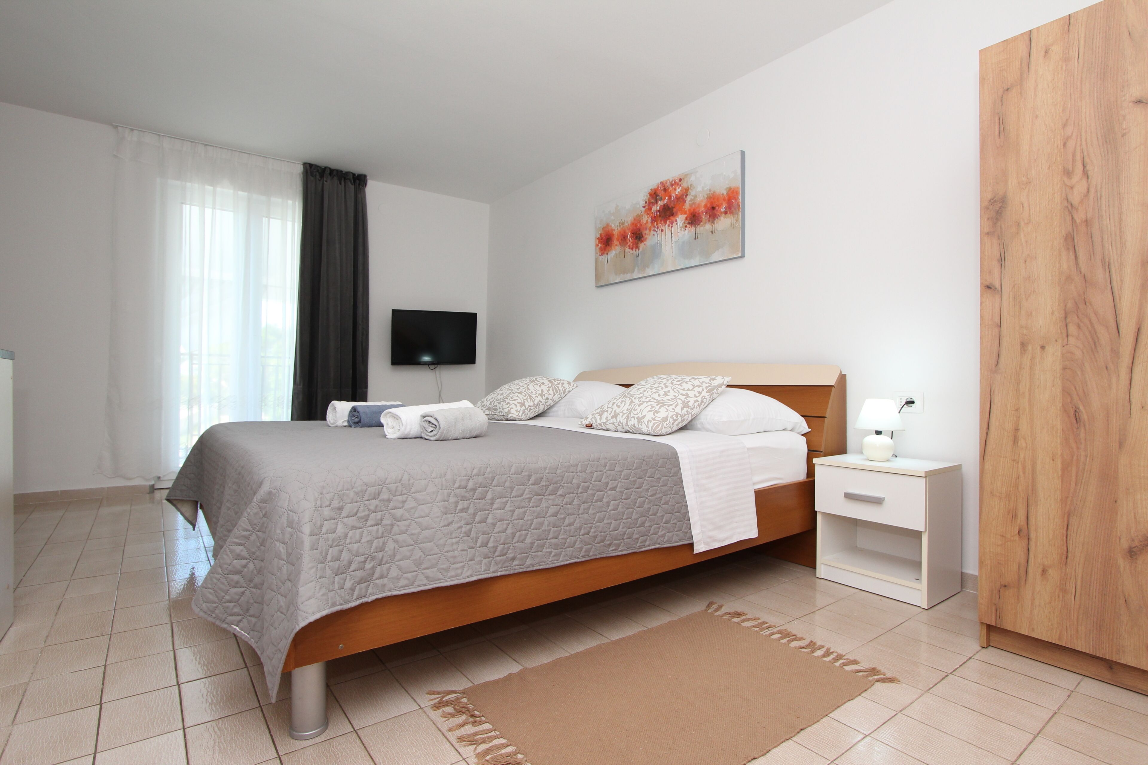 A bedroom with a bed, a bedside table with a lamp, a TV on the wall, and a painting.