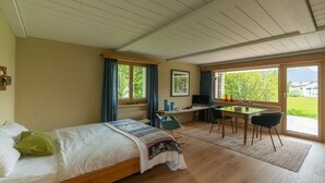 A bedroom with a bed, a desk, and a chair, a window with curtains, and a view of greenery outside.
