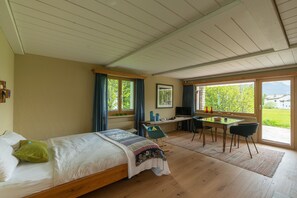 Ein Schlafzimmer mit Bett, Schreibtisch und Stuhl, einem Fenster mit Vorhängen und Blick ins Grüne.
