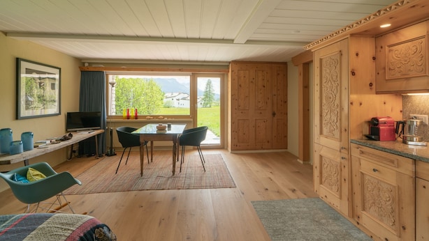 A modern kitchen with wooden cabinets, a dining area with a table and chairs, a television, and a large window with a view of greenery.