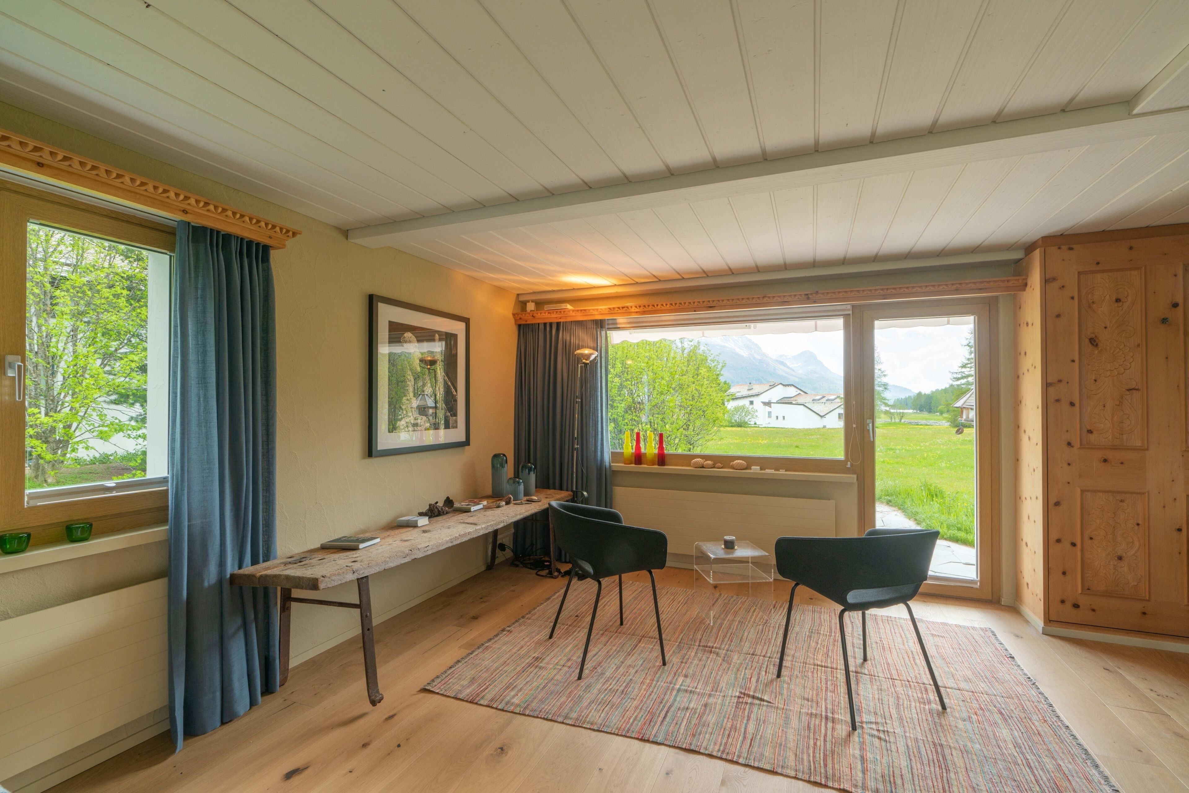 Een kamer met een houten plafond, een groot raam met gordijnen, een houten tafel, twee zwarte stoelen en een vloerkleed.
