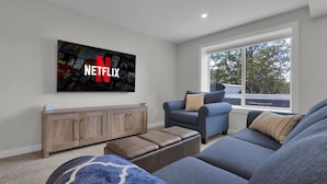 Un salon avec un grand téléviseur fixé au mur, sur lequel apparaît le logo de Netflix, un canapé bleu, un meuble en bois et une fenêtre donnant sur des arbres.