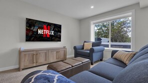 Una sala de estar con una televisión grande montada en la pared que muestra el logotipo de Netflix, un sofá azul, un mueble de madera y una ventana con vistas a árboles.