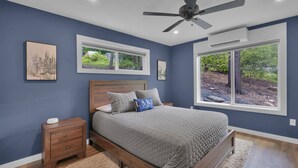 Une chambre à coucher comprenant un lit, des tables de chevet, un ventilateur de plafond et de grandes fenêtres donnant sur un paysage verdoyant.