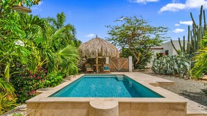 Piscina rodeada por plantas tropicais, uma construção com telhado de palha e cactos.